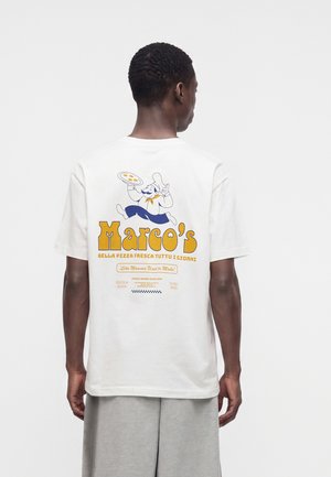 Homme portant un t-shirt blanc avec un graphisme coloré du chef de pizza Marco et un texte au dos, debout devant un fond uni.