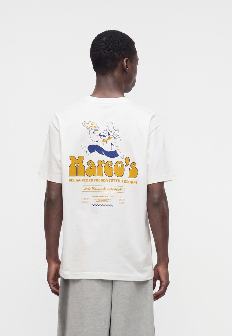 Άνδρας που φοράει λευκό t-shirt με πολύχρωμο γραφικό σεφ πίτσας Marco και κείμενο στην πλάτη, στέκεται μπροστά σε απλό φόντο.