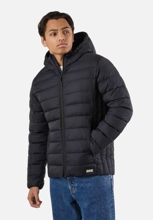 Ung mand iført en sort quiltet hættetrøjejakke med lynlås, hænderne i lommerne, kombineret med blå jeans, kigger let til siden.