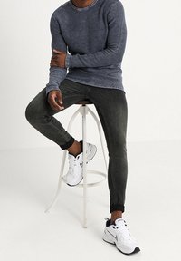 Chemise grise à manches longues côtelée, jean noir slim, et chaussures de sport blanches avec accents noirs. Modèle assis sur un tabouret minimaliste.