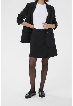 Femme portant un blazer noir, une jupe noire, des collants noirs transparents, une chemise blanche et des mocassins noirs, debout, une main sur la hanche.