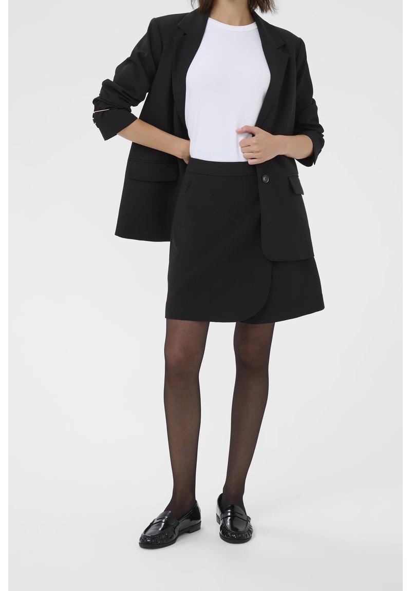 Femme portant un blazer noir, une jupe noire, des collants noirs transparents, une chemise blanche et des mocassins noirs, debout, une main sur la hanche.