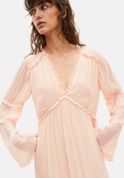 Mujer con cabello ondulado que lleva un vestido plisado de color rosa claro con mangas largas acampanadas y un escote en V profundo, mirando hacia un lado contra un fondo liso.