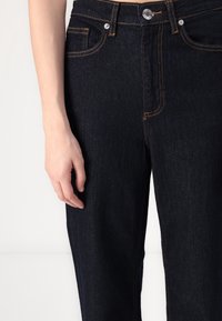 Jeans de mezclilla azul oscuro con costuras visibles en naranja. Cuenta con un diseño clásico de cinco bolsillos y un cierre frontal de botón. Textura suave.