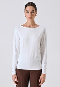 Maglione bianco a maniche lunghe con scollo a barca, texture a coste e dettagli con bottoni sui polsini, abbinato a pantaloni marroni.