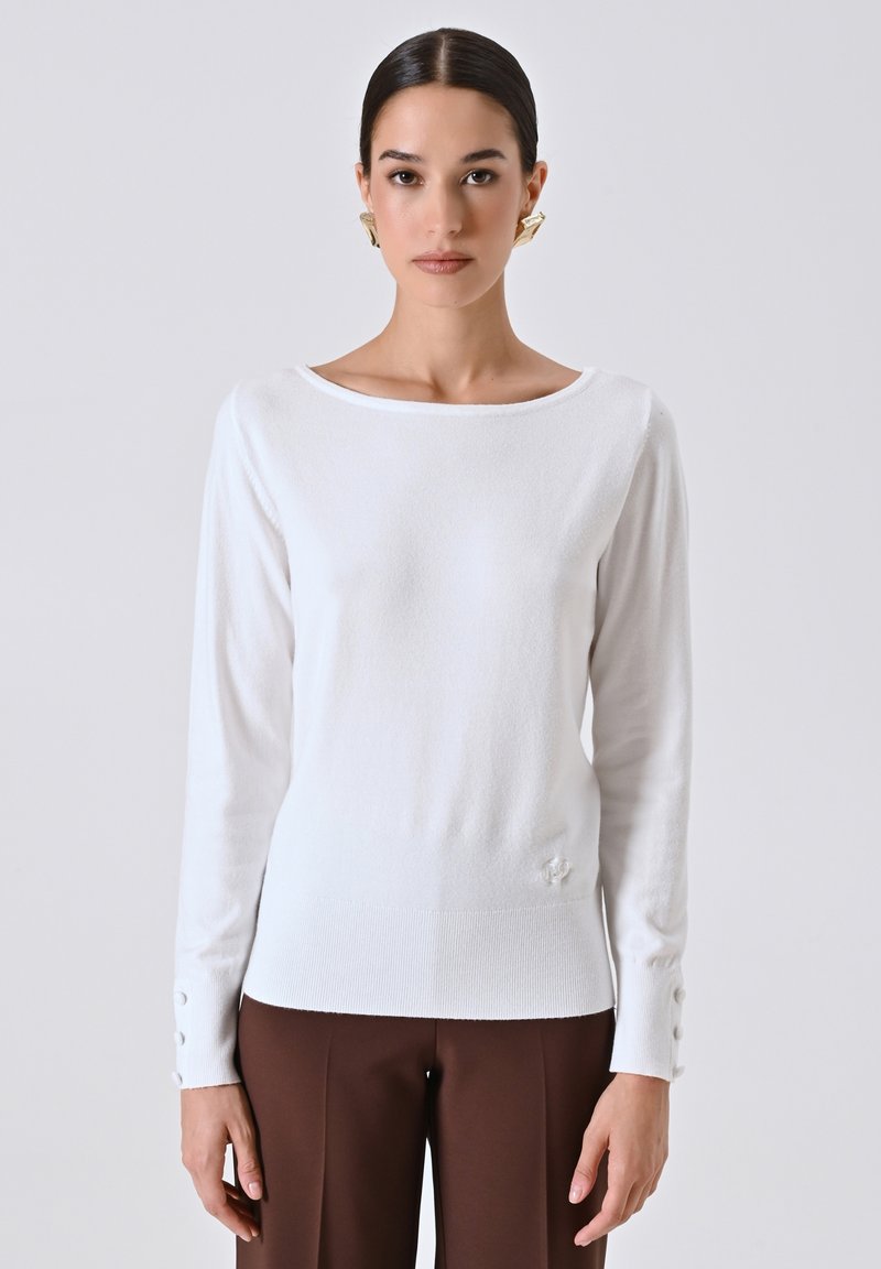 Maglione bianco a maniche lunghe con scollo a barca, texture a coste e dettagli con bottoni sui polsini, abbinato a pantaloni marroni.