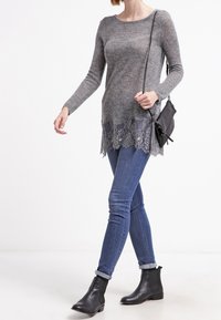 Pull en maille gris avec un ourlet en dentelle, associé à un jean skinny bleu et des bottines noires. La tenue comprend un petit sac en bandoulière noir minimaliste.
