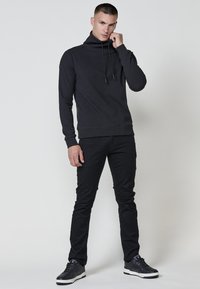 Maglione nero a maniche lunghe con colletto alto e lacci, in tessuto testurizzato. Abbinato a pantaloni neri aderenti e scarpe sportive scure.