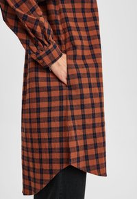 Vestito camicia a scacchi marrone e blu navy con maniche lunghe, orlo arrotondato e tasca laterale, realizzato in tessuto morbido e testurizzato.