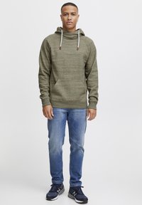 Sudadera verde oliva con acabado texturizado, cuello con cordón, bolsillo frontal, combinada con jeans azules y zapatillas deportivas negras.