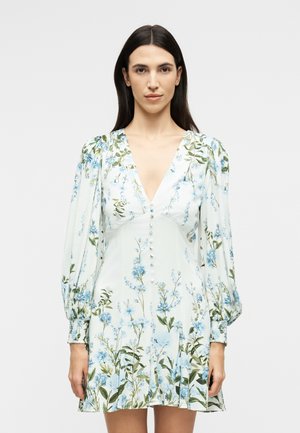 Hosszú sötét hajú nő fehér, rövid, virágmintás ruhában, kék virágokkal, V-nyakkivágással, fodros hosszú ujjakkal és gombos rátétekkel.