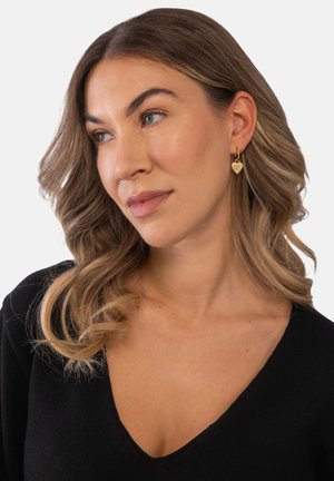 Frau mit welligem hellbraunem Haar, die goldene Ohrringe in Herzform und ein schwarzes V-Ausschnitt-Oberteil trägt, leicht nach links blickend.