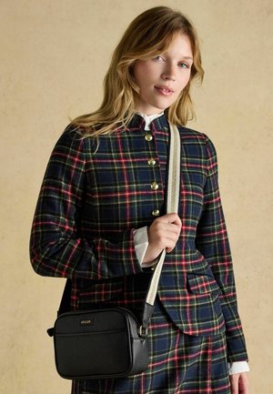 Frau trägt eine marineblaue kariertes Jacke mit goldenen Knöpfen, hält eine schwarze Umhängetasche mit weißem Riemen vor einem beigen Hintergrund.