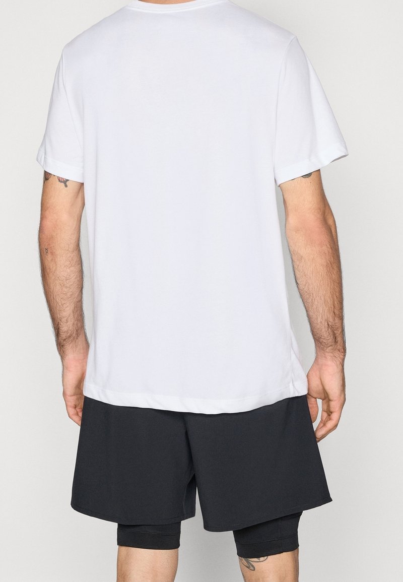 Homme portant un t-shirt blanc uni à manches courtes et un short noir superposé, de dos devant un fond uni.