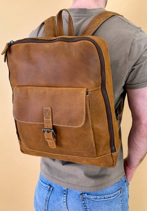 Brown leather backpack with zip closure, front flap pocket, adjustable straps, and stitched accents, displayed on a person wearing a t-shirt.