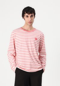 Ilga rankovė marškiniai rožinės ir baltos juostelėmis, su apvaliame iškirpime ir mažuoju raudonu gėlių simboliu krūtinėje. Minkšta medvilnė.