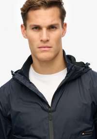 Navy windbreaker jacket with a zippered front, hood, and adjustable toggles. Made from smooth, lightweight material; features subtle branding.