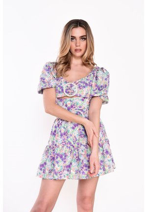 Kobieta w krótkiej sukience w kwiaty z bufkami i detalami z perełkami na klamrach, pozująca z rękami skrzyżowanymi na tle białego tła.