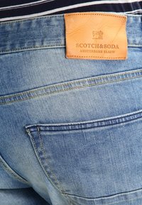 Ljusblå denimjeans med en läderlapp märkt "SCOTCH&SODA" vid midjan, med bakfickor och sydda detaljer.