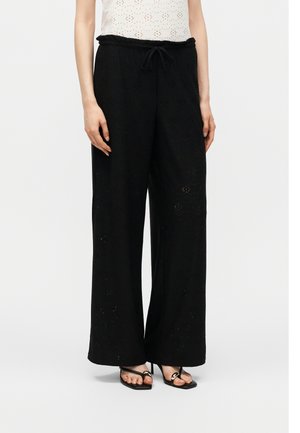 Άτομο που φοράει μαύρο παντελόνι με φαρδιά πόδια και φλοράλ σχέδιο με τρυπητά μοτίβα και μαύρα σανδάλια με τακούνι, στέκεται μπροστά από μια απλή φόντο.