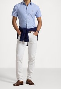 Světlomodrá krátká košile na knoflíky, bílé slim fit kalhoty, tmavě modrý svetr uvázaný kolem pasu a hnědé mokasíny.