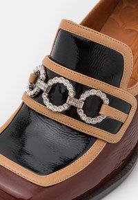 Dessus en cuir verni noir avec des accents beiges et une boucle décorative ornée de strass. Bout arrondi et semelle bordeaux brillante.