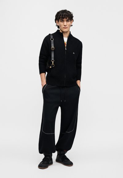 Μαύρο πλεκτό πουλόβερ με φερμουάρ και ανάγλυφη υφή, συνδυασμένο με χαλαρά μαύρα παντελόνια με αντίθετη ραφή και ζώνη με κορδόνι.
