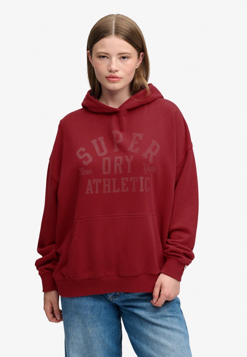 Młoda kobieta z prostymi jasnobrązowymi włosami nosi luźną czerwoną bluzę z kapturem z napisem "Super Dry Athletic" i niebieskie dżinsy na białym tle.