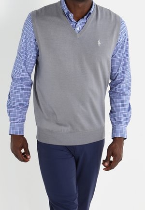 Mann trägt einen grauen ärmellosen Pullover über einem blau karierten Hemd und dunkelblaue Hosen, geht vorwärts vor einfachem Hintergrund.