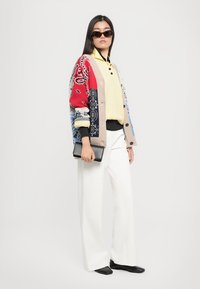 Femme portant des lunettes de soleil, un gilet à motifs, un pull jaune, un pantalon blanc, tenant une pochette noire, se tenant devant un fond clair.