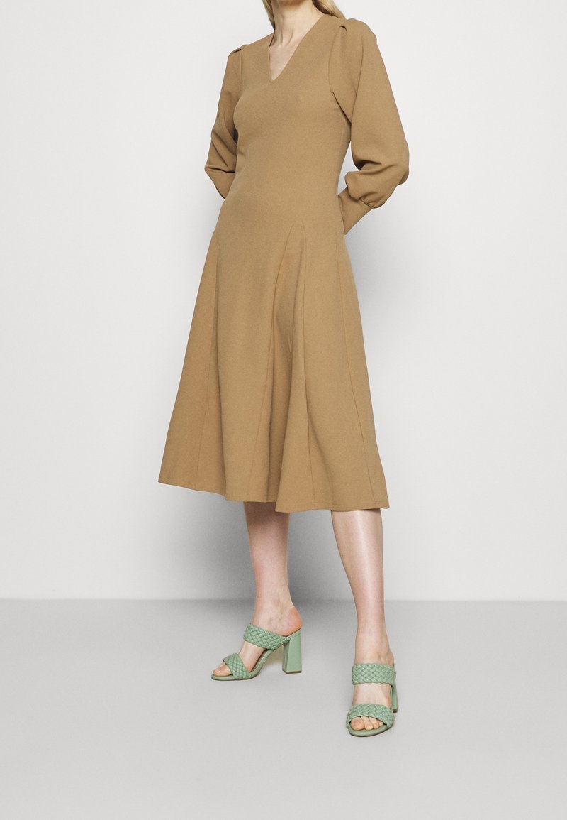 Robe marron, longueur genoux, avec un décolleté en V et des manches bouffantes. Portée avec des sandales à talons blocs tressées vertes. Tissu lisse, design simple.