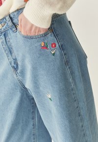 Jeans in denim azzurro chiaro con fiori ricamati in rosso, arancione e verde sulla tasca frontale. Presentano una vestibilità dritta e cuciture visibili.