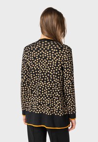 Svart cardigan med ett prickmönster i beige. Långa ärmar, rund nederkant med en gul trim. Mjuk textur, avslappnad passform. Bakvy.