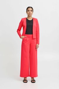 Heller pink maßgeschneiderter Blazer und weit geschnittene Hose, kombiniert mit einem schwarzen gerippten Oberteil. Glatter Stoff mit Taschen an der Hose, schwarze Sandalen.