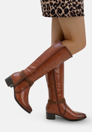 Bottes marron jusqu'aux genoux en cuir lisse, avec une sangle ornée de matériel décoratif et un talon modeste. Jupe en imprimé animal texturé.
