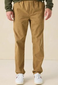 Homme portant un pantalon en velours côtelé beige et des baskets blanches, se tenant devant un fond clair uniforme.