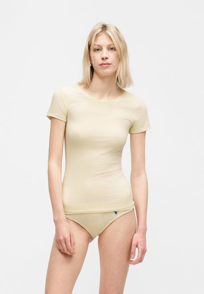Jeune femme aux cheveux blonds portant un t-shirt beige ajusté et des sous-vêtements assortis, debout devant un fond clair uni.