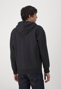 Sudadera negra con bolsillo canguro, puños y dobladillo de canalé; combinada con jeans oscuros. La tela parece suave y ligeramente texturizada.