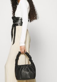 Donna con capelli ricci che indossa un top bianco a maniche lunghe e una cintura nera con borchie, mentre tiene in mano una borsa nera con manico arricciato.