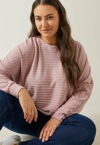 Helles rosa Sweatshirt mit vertikaler Rippstruktur und langen, leicht gepufften Ärmeln. Kombiniert mit dunkelblauen Jeans und weißen Sneakers.