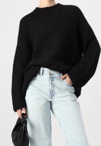 Musta, ylisuuressa neulepuserossa on pyöreä kaula-aukko ja pitkät hihat, ja se on yhdistetty vaaleansinihin, l öysiin farkkuihin sekä mustaan laukkuun.