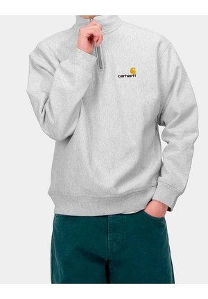 Persoon in lichtgrijze Carhartt kwart-rits sweatshirt met logo op de borst, hand die de rits vasthoudt, donkergroene broek, één hand in de zak.