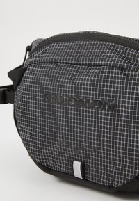 Black fabric waist bag with white grid pattern, featuring a zipper closure and a textured logo. Accent tab in silver at the base.
