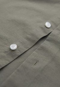 Camicia verde oliva realizzata in tessuto testurizzato, dotata di bottoni bianchi, una placca e un sottile dettaglio di cucitura vicino all'area dei bottoni.
