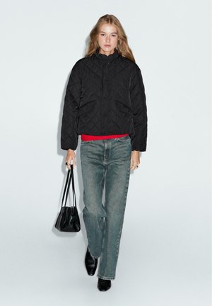 Schwarze gesteppte Jacke mit hohem Kragen, Fronttaschen, kombiniert mit rohem Hemd, hellblauen Jeans, schwarzen Schuhen und einer kleinen schwarzen Tasche.