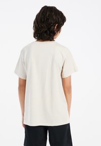 Camiseta de algodón beige de manga corta, con cuello redondo y dobladillo recto. La parte trasera es lisa, sin etiquetas ni gráficos visibles.