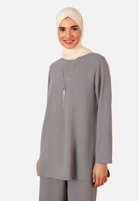 Light gray long-sleeve tunic with a loose fit, made of textured fabric. A beige scarf and a long silver pendant necklace complement the outfit.