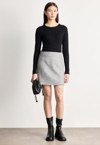 Νεαρή γυναίκα με μακριά σκουρόχρωμα μαλλιά, ντυμένη με μαύρο μακρυμάνικο τοπ, γκρι υφασμάτινη μίνι φούστα και μαύρα μπότες, κρατώντας μια μικρή μαύρη τσάντα.