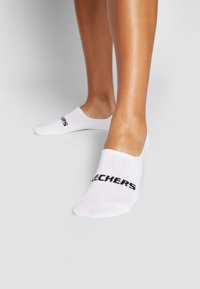 Baltās nesaredzamās zeķes, kas izgatavotas no mīkstās auduma, ar melnu logo tekstu "SKECHERS" sānos. Dizains ir zems, atsedzot ādu virs potītes.