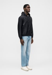 Μαύρο ελαφρύ μπουφάν με κουκούλα και υφή σε σχέδιο, συνδυασμένο με ανοιχτό μπλε τζιν παντελόνι ίσιας γραμμής και λευκά αθλητικά παπούτσια.